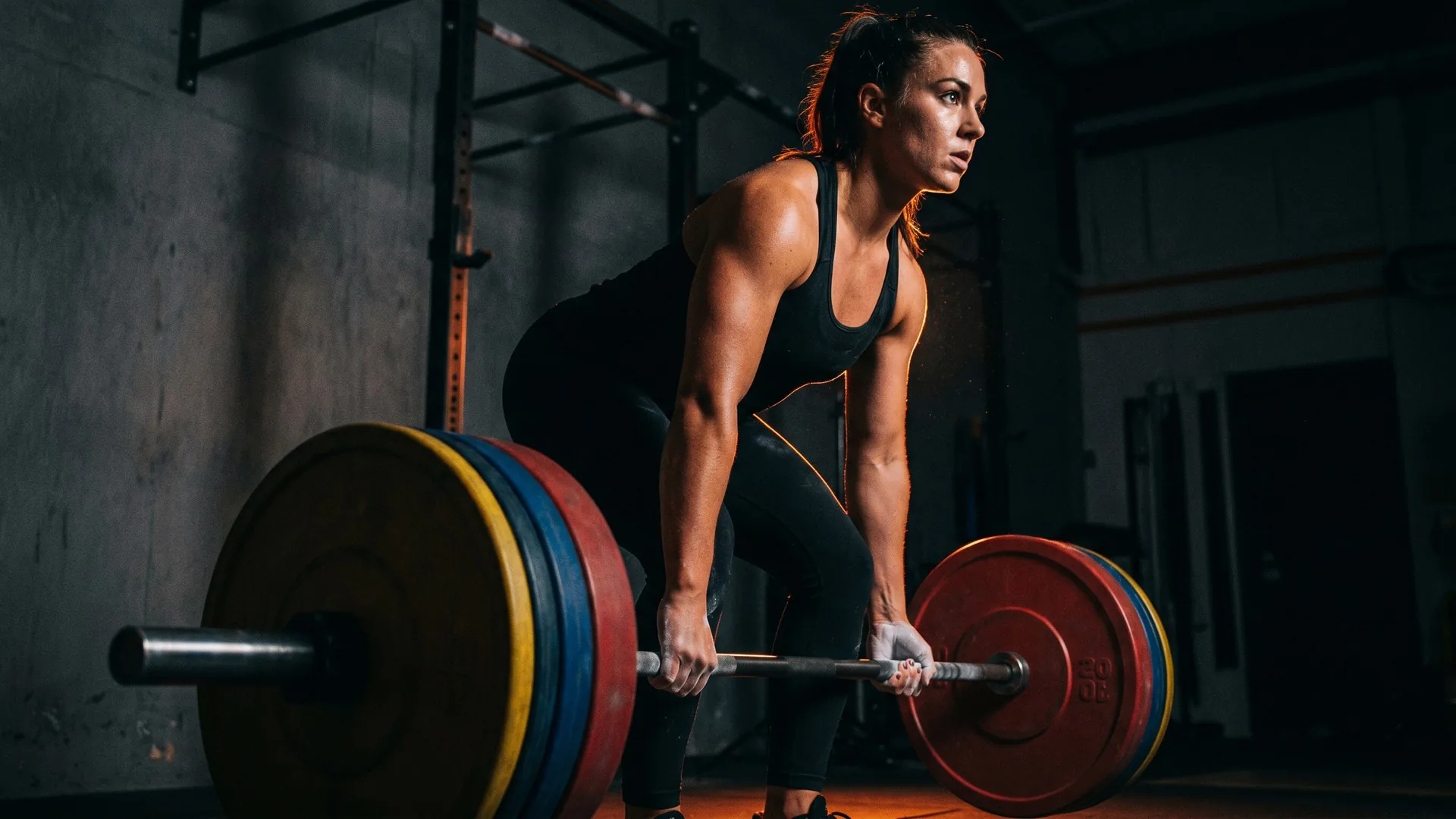 Loaded barbell resting on the floor before a deadlift in a dimly lit gym, Flame Orange rim light catching the plate edges — the iconic deadlift setup